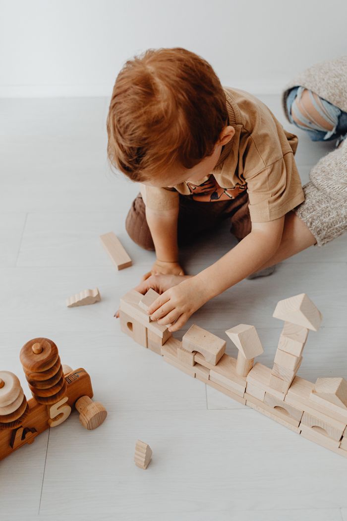 A red-haired toddler builds a row of natural wooden blocks and towers on a pale floor beside a wooden toy train.