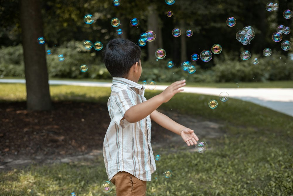 A small boy in a striped shirt, seen from the side, reaches toward dozens of soap bubbles floating in a shady park.