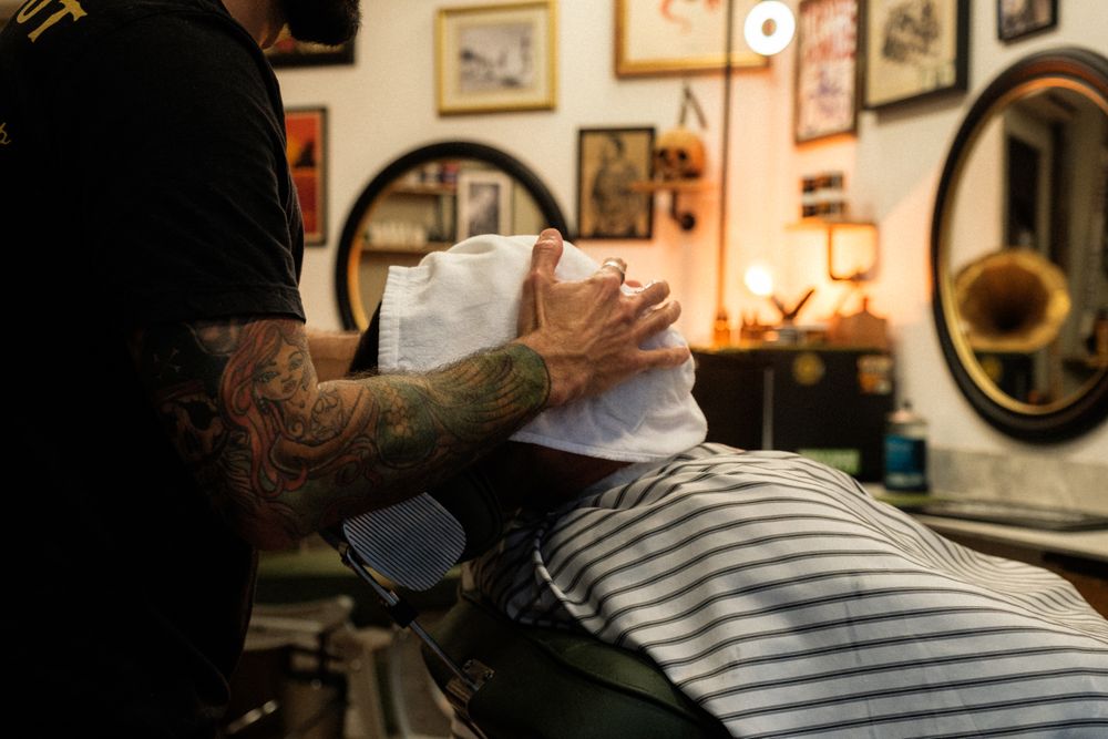 A tattooed barber in a black T-shirt lays a white hot towel over a reclining client's face in a vintage-style barbershop with framed pictures and warm lamps behind.