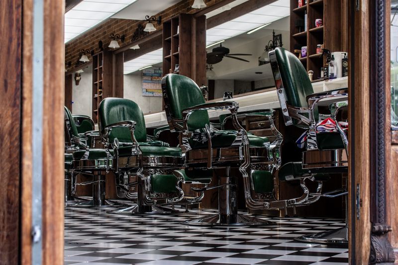 A vintage barbershop seen through a wooden doorway, with a row of green leather chairs with chrome frames on a black and white checkered floor.