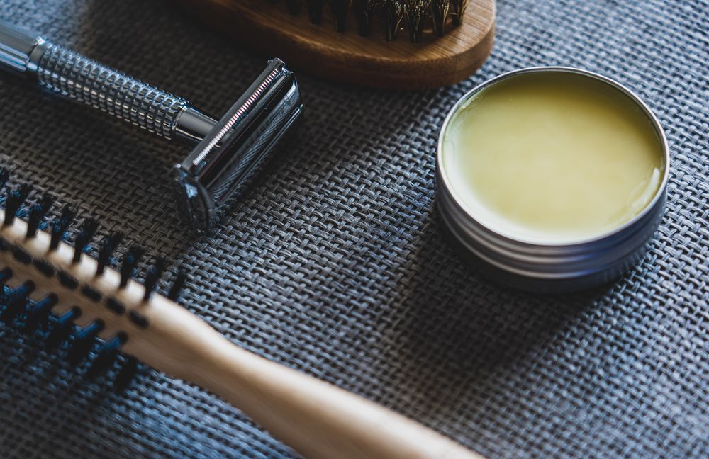 Angled flat lay of an open tin of pale-yellow balm beside a chrome safety razor and two wooden-handled brushes on a grey-blue woven mat.