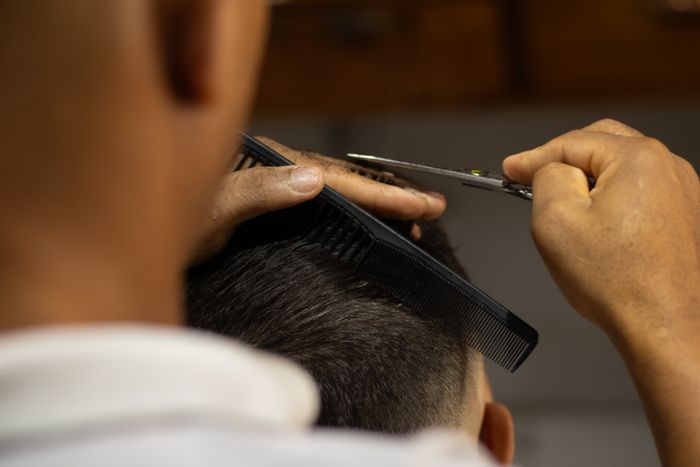 Close-up of hands cutting dark hair with scissors and a black comb, seen from behind.