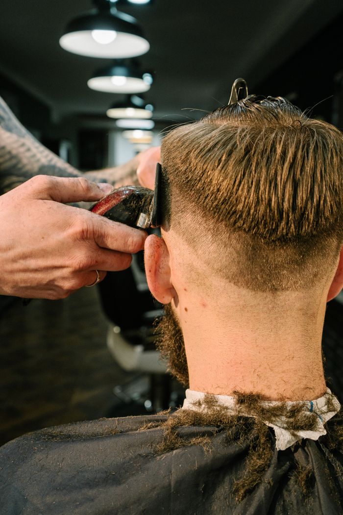 A barber with tattooed forearms trims the back of a seated client's head with an electric clipper and a black guide comb.