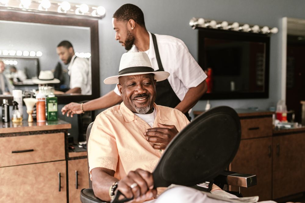 A smiling man in a cream hat and peach shirt looks into a hand mirror in the barber chair while his barber stands behind him in a black apron.