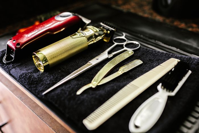 Barber tools laid out on a black towel: a red cordless clipper, a gold trimmer, scissors, a gold straight razor, a cream comb and a white-handled brush.