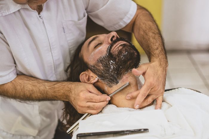 A barber in a white shirt holds a straight razor to the jaw of a reclining bearded client with a towel across his chest.