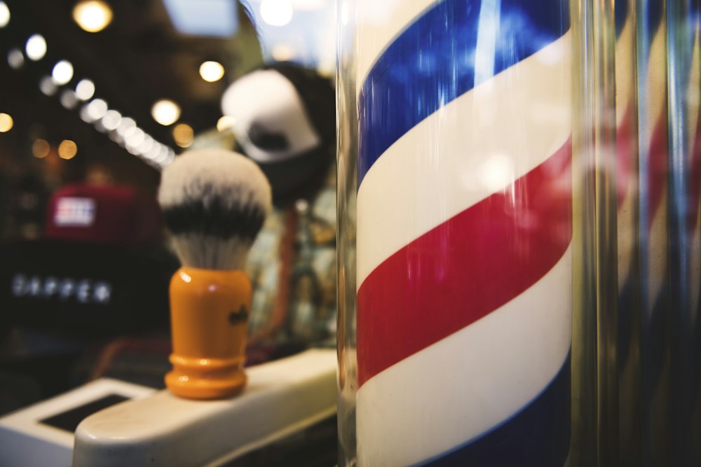 An orange-handled shaving brush with a silvertip knot stands in front of a blurred red, white and blue barber pole.