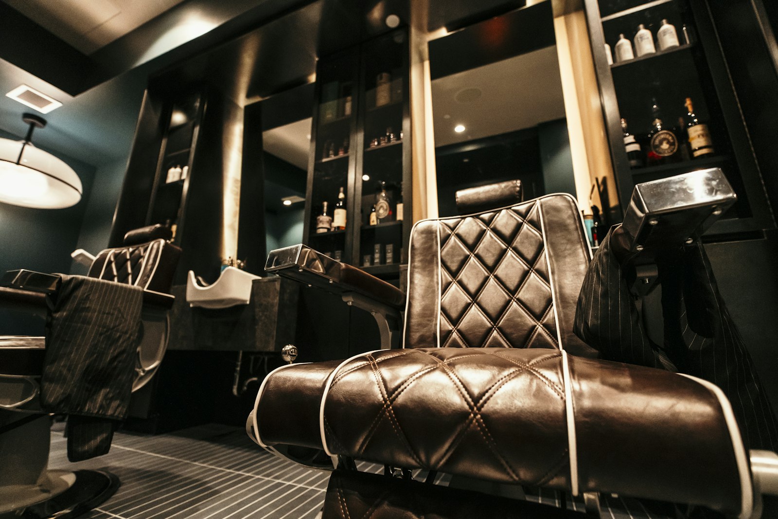 A quilted brown leather barber chair with white piping and chrome armrests in a dark teal barbershop, a second chair, a wash basin and lit bottle shelves behind it.
