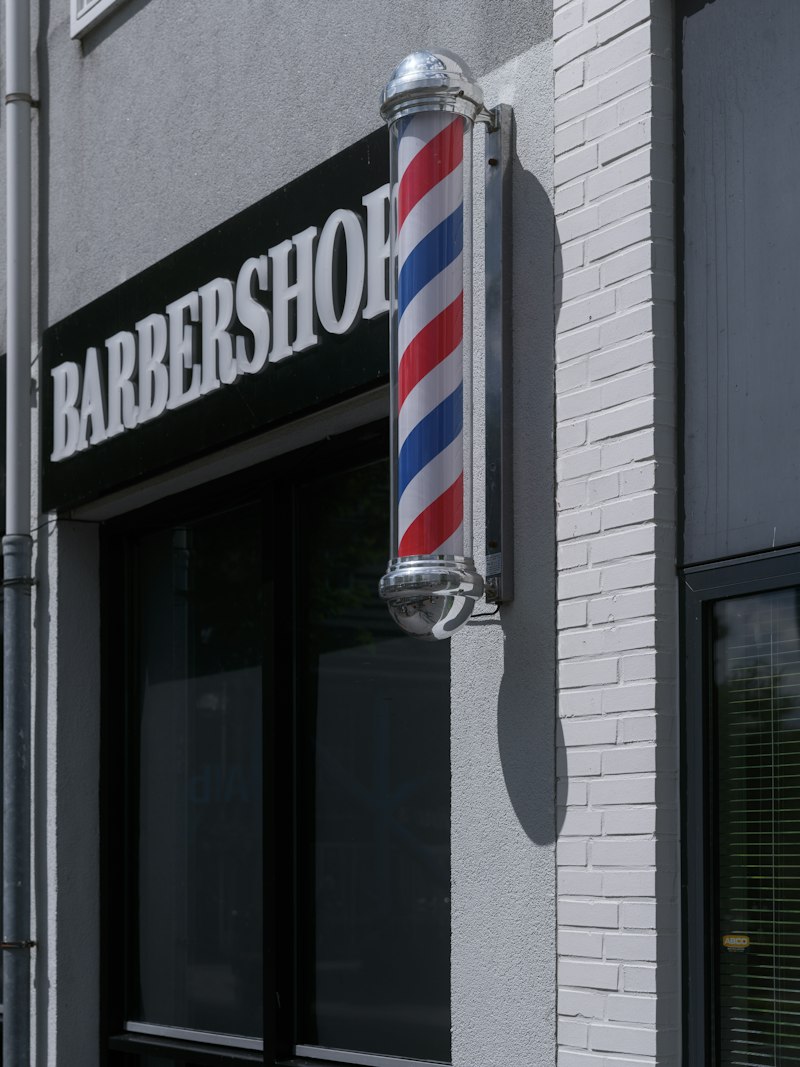 A chrome-capped barber pole mounted on a grey rendered wall beside a white-painted brick pier and a black sign with white BARBERSHOP lettering above a dark shop window.